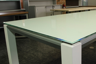 Белый конференц стол из стекла с белыми опорами из искусственного камня. Торец столешницы декорирован алюминиевым профилем